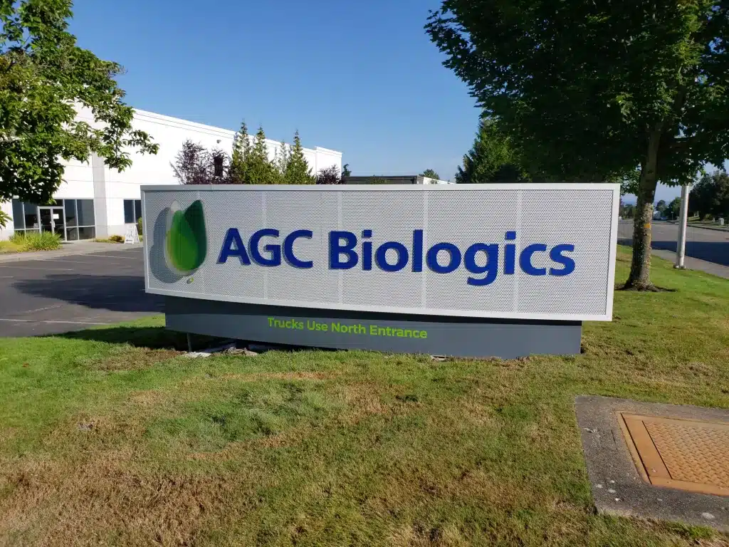 Sign reading "AGC Biologics" with an adjacent logo in a business park, surrounded by grass and trees.