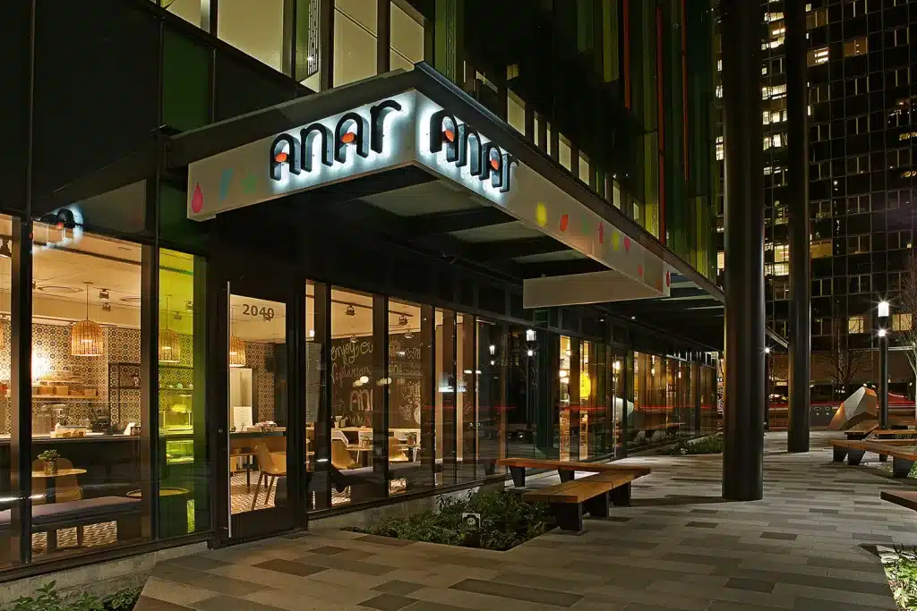 Exterior view of a modern restaurant with glass walls and illuminated signage, located in an urban setting with surrounding buildings.