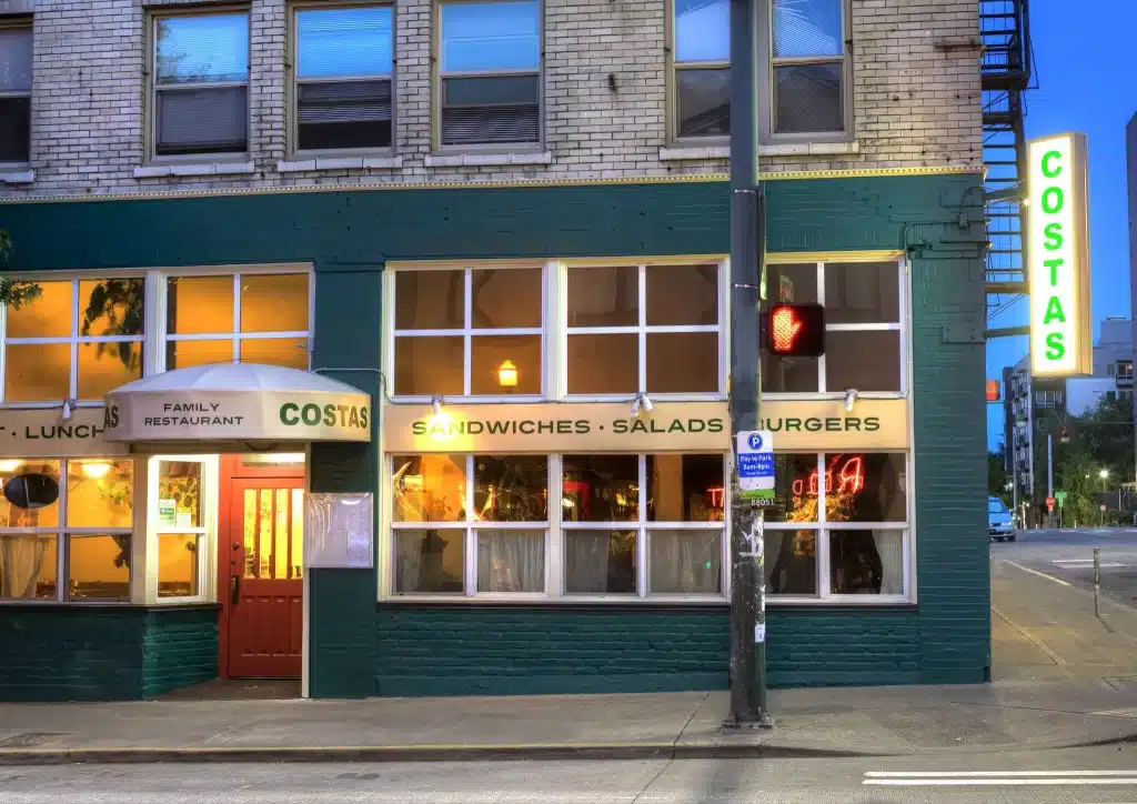 Street view of a corner restaurant with a green and brick exterior. The sign reads "COSTAS" and the windows display "Sandwiches, Salads, Burgers." A traffic light is in the foreground.