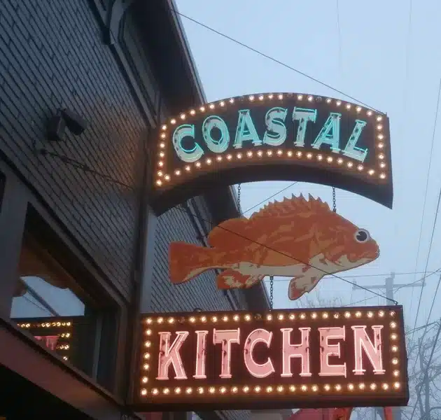 Sign for "Coastal Kitchen" with a lit-up fish graphic, attached to a brick building.