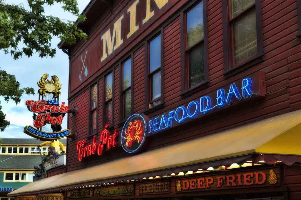 A seafood bar and restaurant with neon signs reading "The Crab Pot" and "Seafood Bar" on a rustic building's exterior.