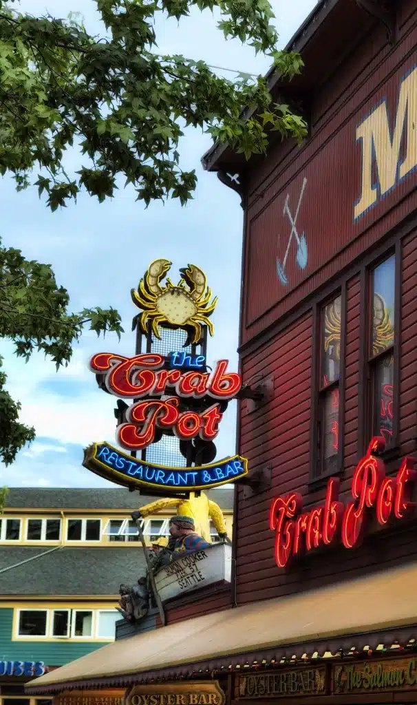 Sign for "The Crab Pot Restaurant & Bar" with a crab illustration, attached to a wooden building exterior. Trees partially frame the scene.