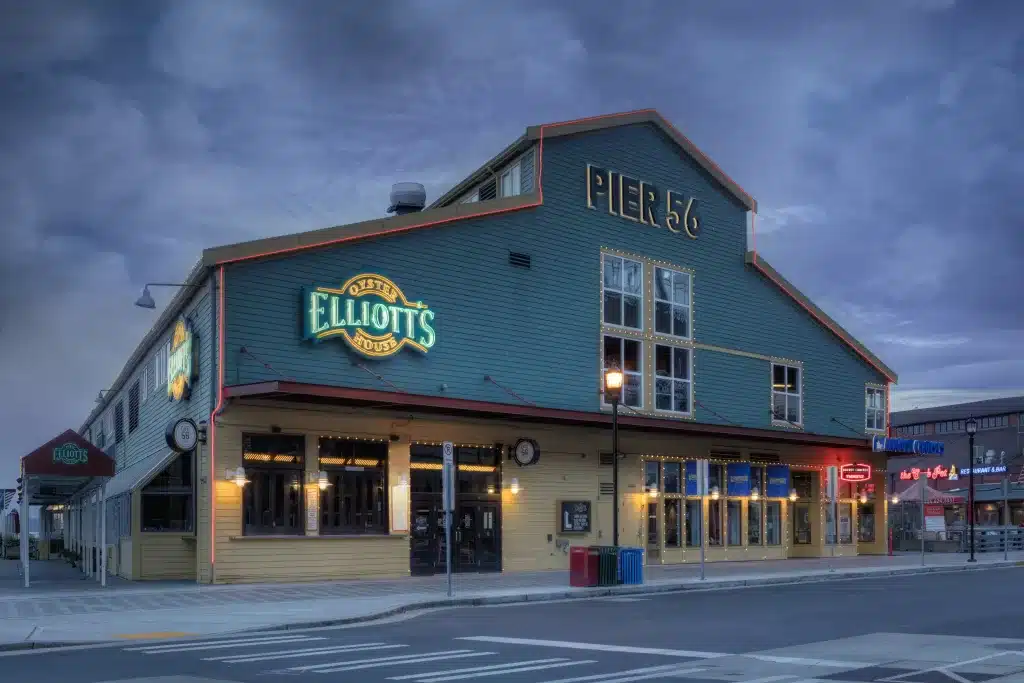 Street view of a two-story building with "Elliott's" neon sign and "Pier 56" on the roof against a cloudy sky.