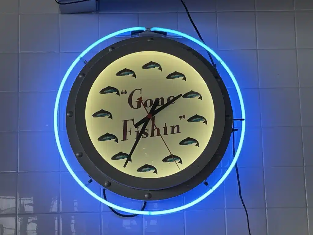 A round neon clock with a blue outer ring, featuring fish graphics and the words "Gone Fishin'" on a tiled wall.