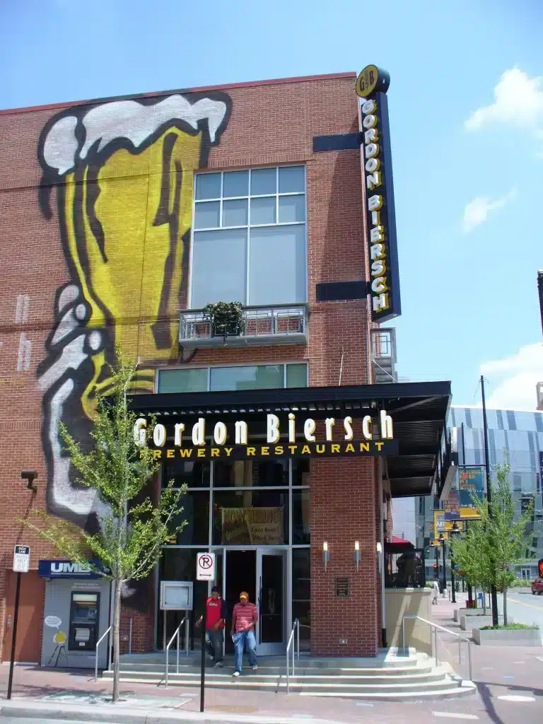 Front view of Gordon Biersch Brewery Restaurant with a mural of a beer glass on the brick wall. Two people are walking towards the entrance.