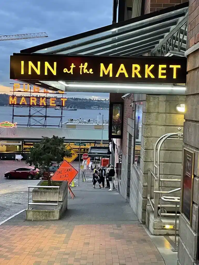 Sign reading "Inn at the Market" with a view of Pike Place Market and Elliott Bay in the background. Brick pathway leads down to a street below.