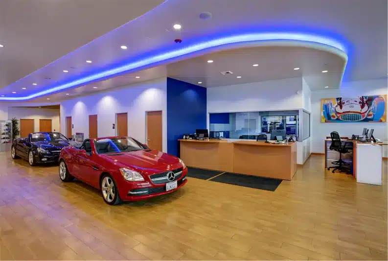 Car dealership showroom with two cars on display, including a red convertible. Reception desk with office chairs and computers in the background, and bright overhead lighting.