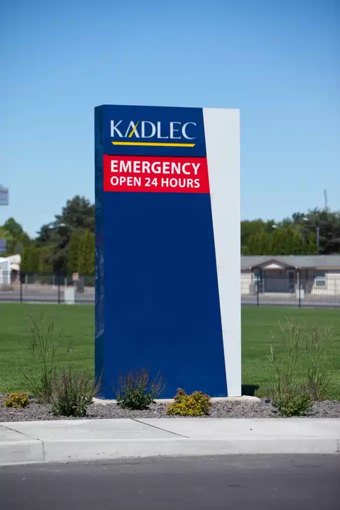 Blue and white sign with "Kadlec Emergency Open 24 Hours" text, standing on a grassy area with visible plants.