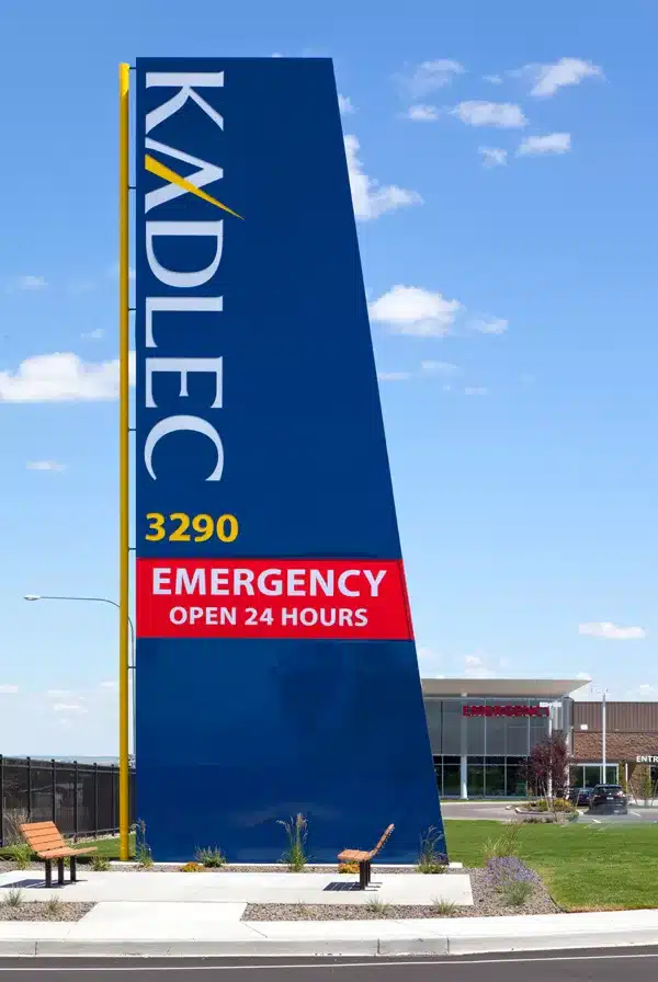 Sign displaying "Kadlec 3290 Emergency Open 24 Hours" in front of a hospital building under a clear blue sky.