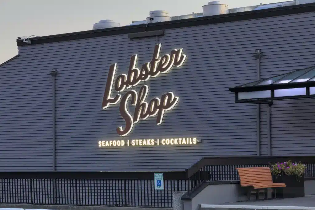 Building with "Lobster Shop" sign in cursive, offering seafood, steaks, and cocktails. There is a railing, a bench, and a handicap parking sign in front.