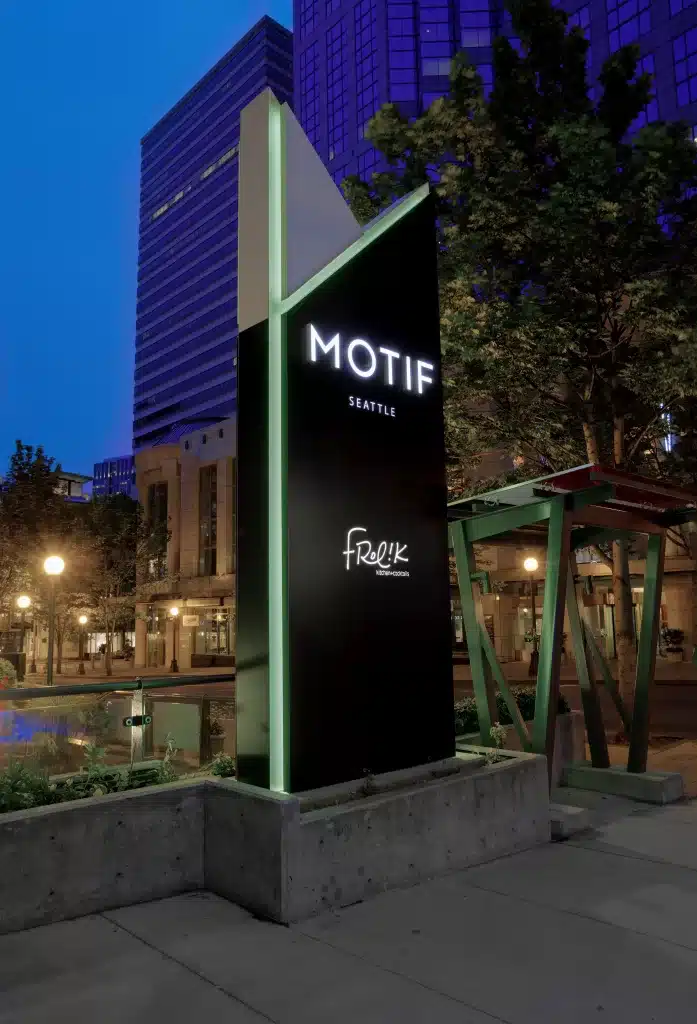 Sign for Motif Seattle hotel at dusk, with Frolik Kitchen + Cocktails mentioned. Urban setting with modern buildings and trees in the background.