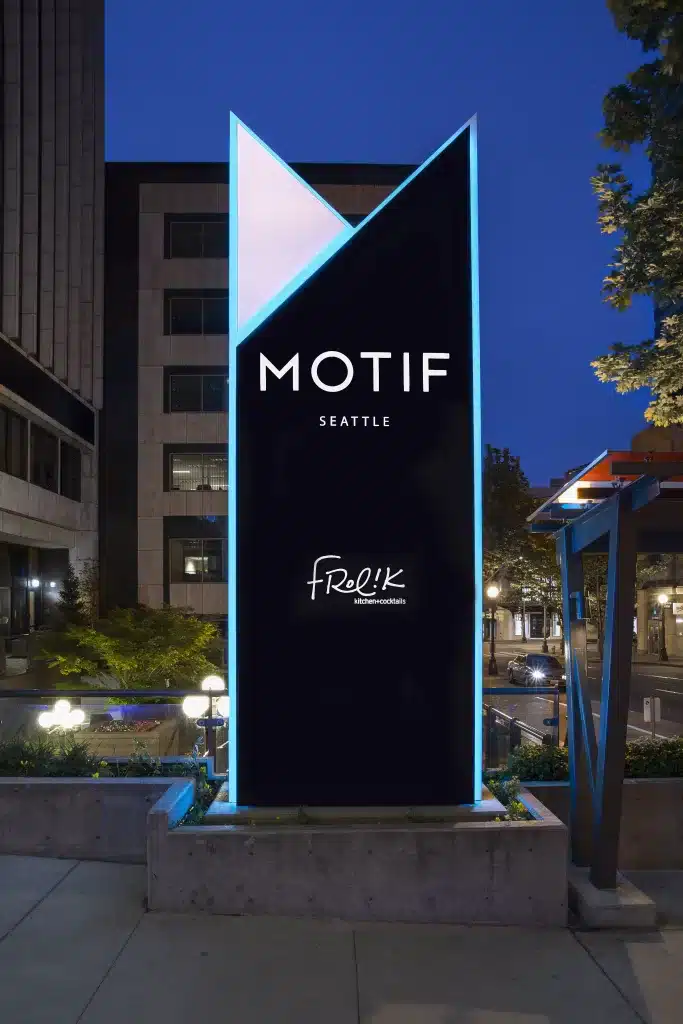 Sign for Motif Seattle and Frolik Kitchen+Cocktails, with a modern geometric design, lit up at night against an urban backdrop.