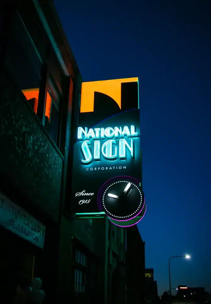 Neon sign for "National Sign Corporation" with a clock below it on a building facade at dusk.