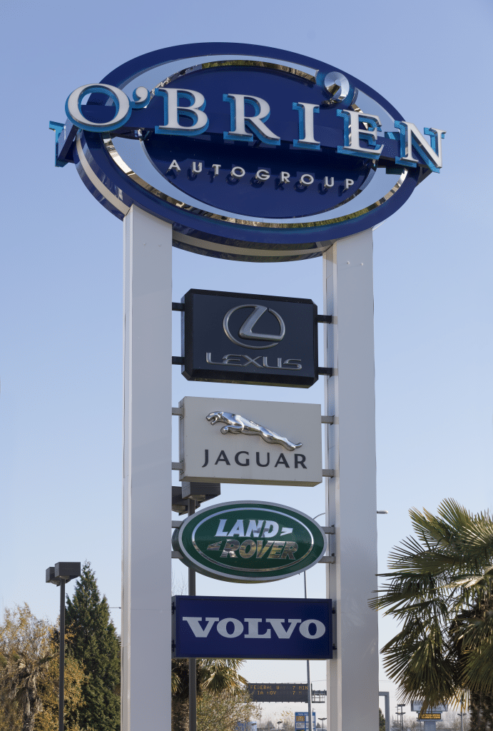 Car dealership sign featuring O'Brien Autogroup, Lexus, Jaguar, Land Rover, and Volvo logos, set against a clear blue sky.