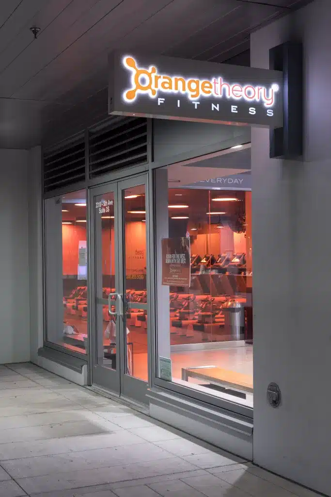 Exterior view of an Orangetheory Fitness studio with illuminated sign and orange-tinted interior.