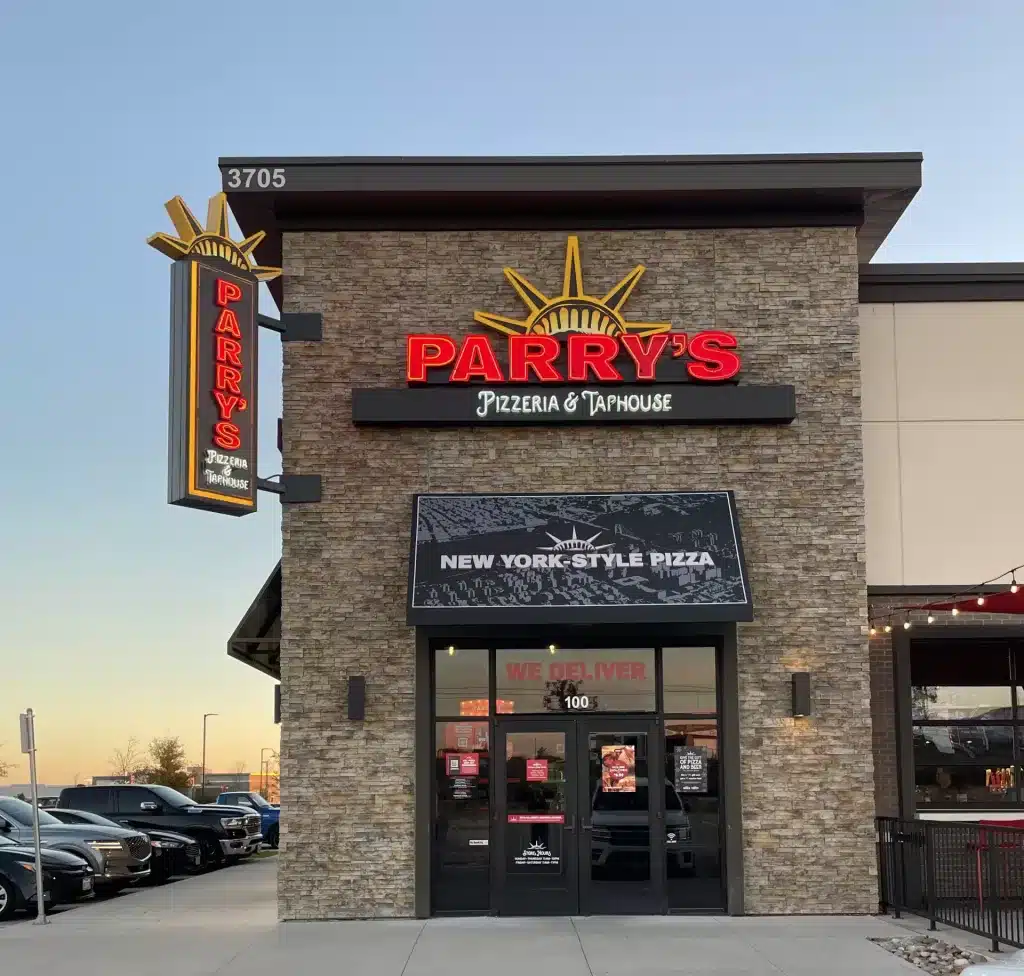 Exterior view of Parry's Pizzeria & Taphouse with a sign advertising New York-style pizza. The building is made of stone with large glass windows. Cars are parked nearby.