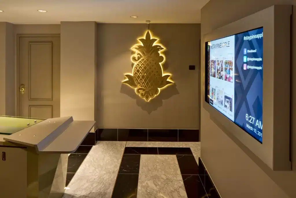 Hallway with illuminated pineapple wall art and a large digital display showing social media feeds and the time, 8:27 AM.