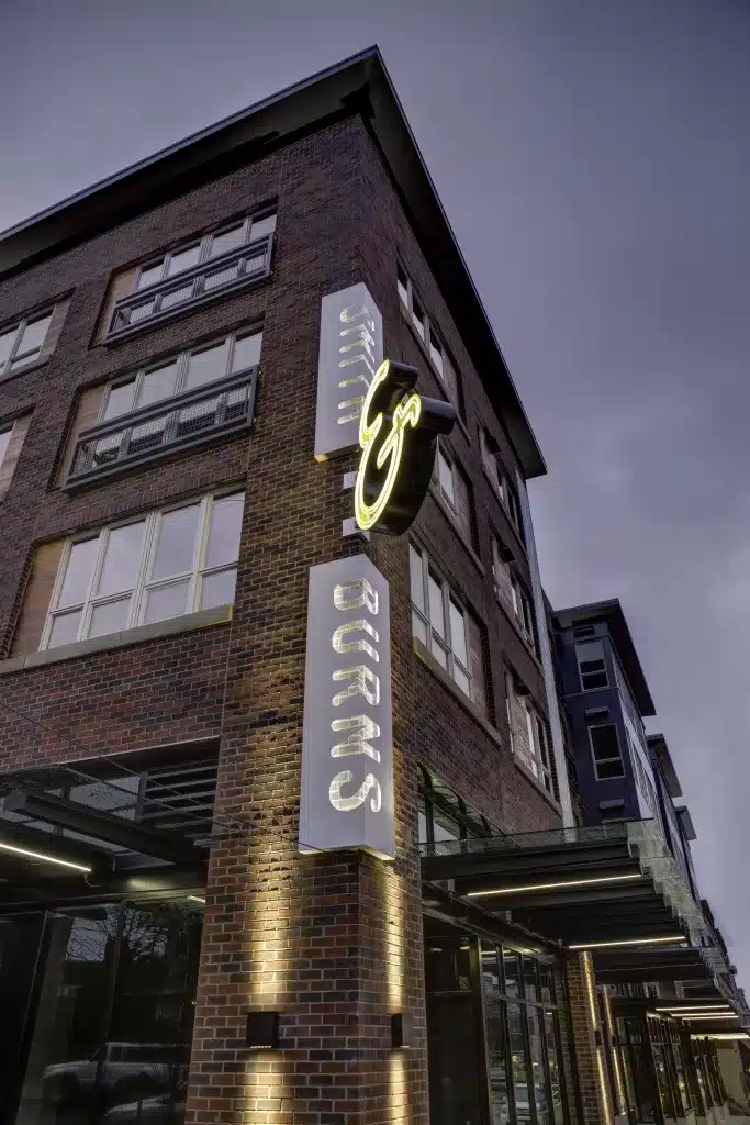 A tall brick building with a neon sign reading "Burns &" at dusk, featuring large windows and exterior lights.