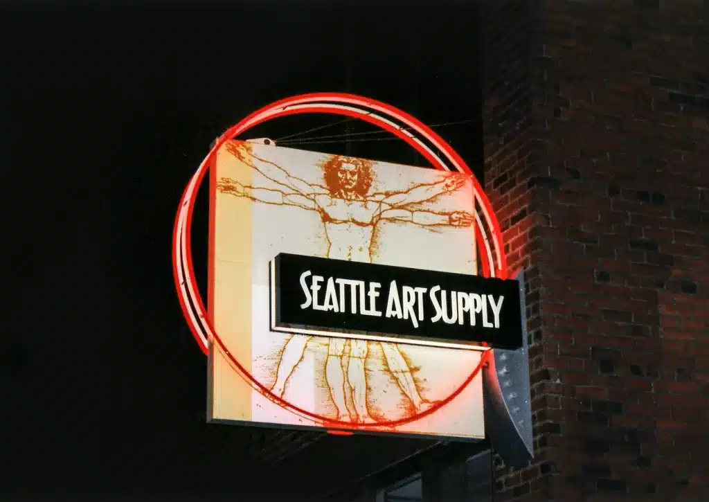 Illuminated sign for Seattle Art Supply featuring a Vitruvian Man design within a red neon circle on a brick building at night.