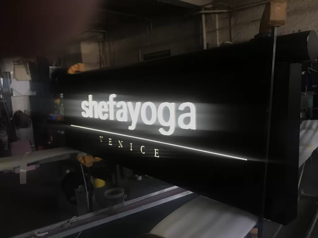 Illuminated sign reading "shefayoga VENICE" in a dimly lit environment.