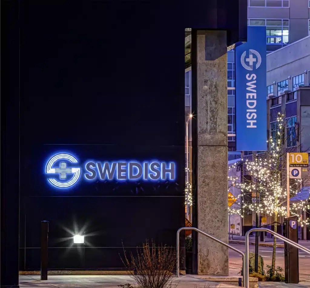 Neon sign for "Swedish" on a building at dusk, with nearby decorative lights and street signs visible.