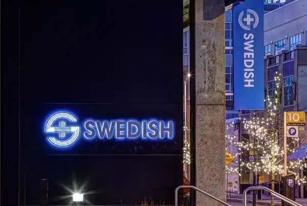 Neon sign for "Swedish" on a building at dusk, with nearby decorative lights and street signs visible.