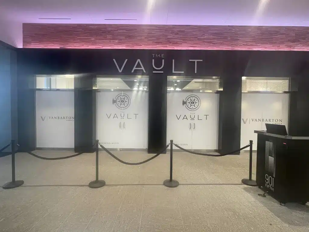 A storefront with black and white signage reads "The Vault" and "Vanbarton." Three covered windows are labeled "Coming Soon." Velvet ropes are set in front.