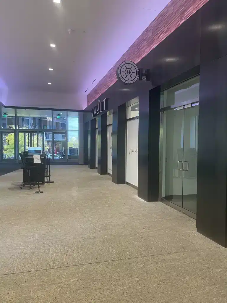 A modern, empty hallway with dark walls and a sign labeled "Vault." Glass doors and a reception desk are present, with natural light coming through large windows.