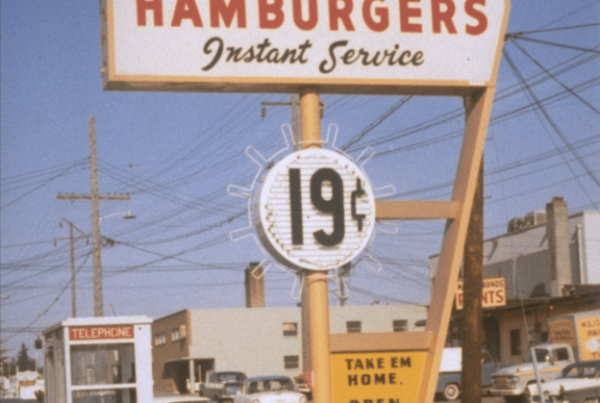 Vintage hamburger sign advertising "Instant Service" and hamburgers for 19 cents. A small notice reads "Take Em Home, Open 11 A.M. - 2 A.M." Surrounding area shows cars and a phone booth.