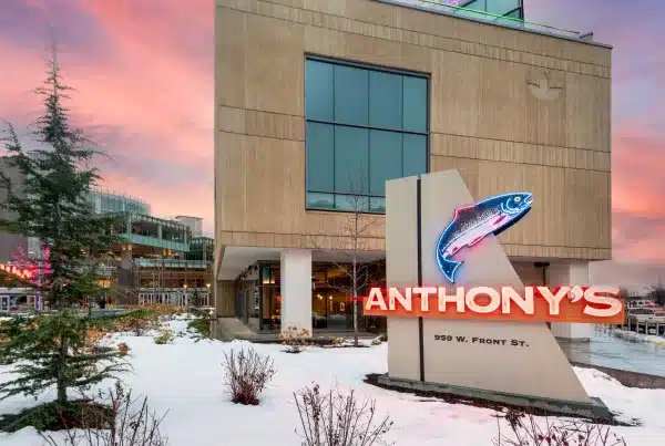 Front view of a building at sunset with a neon "Anthony's" sign and a fish graphic. Snow is on the ground and trees are in the foreground.