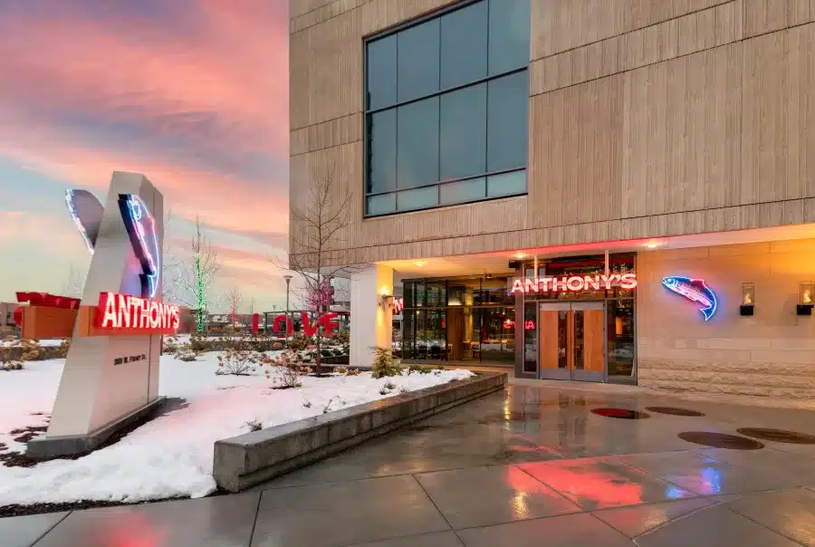 Entrance of Anthony's restaurant with neon signs, set against a modern building. Snow on the ground and a colorful sunset sky in the background.