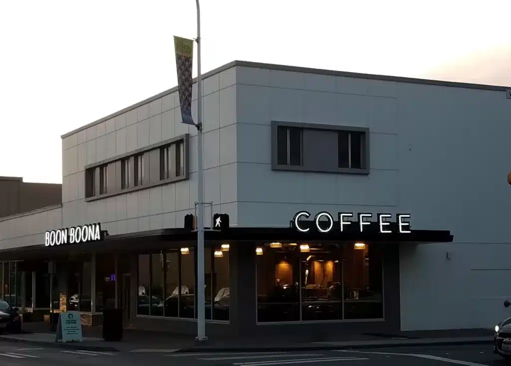 A modern corner coffee shop with a matte exterior, large glass windows, and illuminated signage reading "Boon Boona COFFEE.