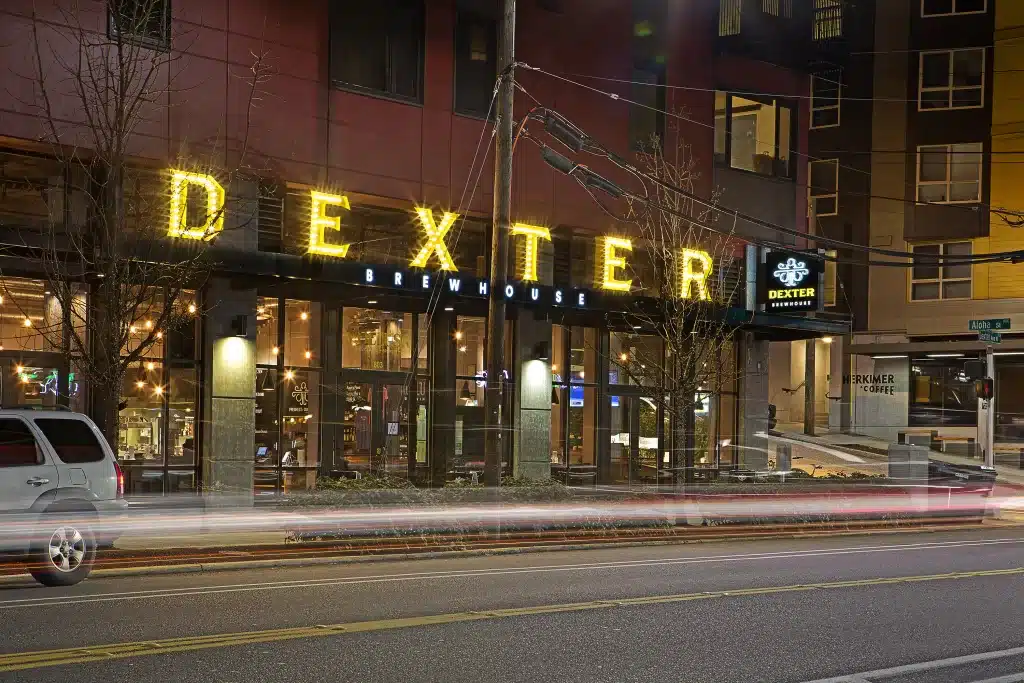 Night view of Dexter Brewhouse with illuminated signs. Motion blur from passing cars on the street in front.