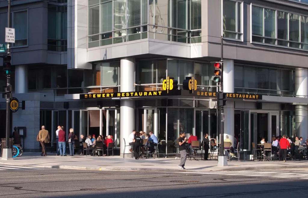 Street view of a brewery restaurant with outdoor seating. People are dining under a modern building with large windows. A pedestrian is crossing the street in the foreground.