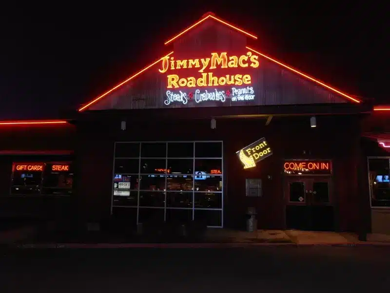 Exterior of Jimmy Mac's Roadhouse at night, featuring neon signs for steaks, gift cards, and a front door entrance.