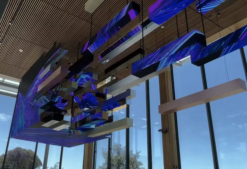 Ceiling installation featuring suspended rectangular elements with purple and blue lighting, against large glass windows and blue sky.