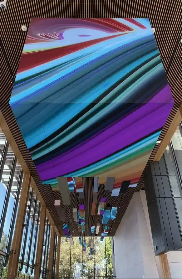 Colorful abstract digital art displayed on a ceiling installation, featuring vibrant swirling patterns in blue, purple, and green, with wooden ceiling panels and large windows in the background.