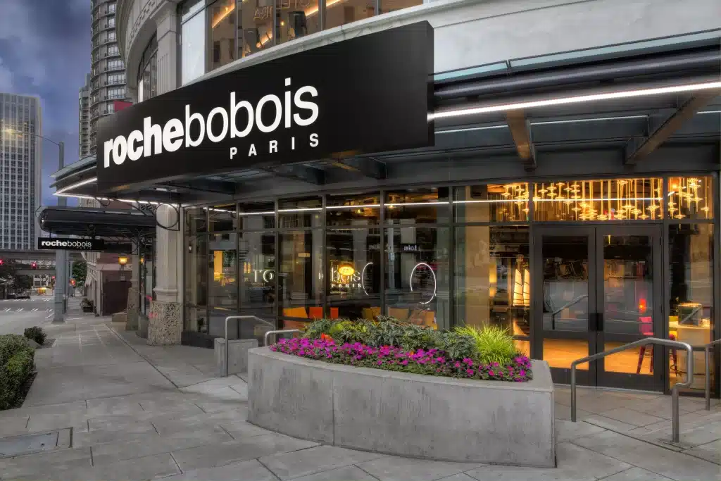 Storefront of Roche Bobois Paris with a black and white sign, surrounded by urban architecture and floral planters.