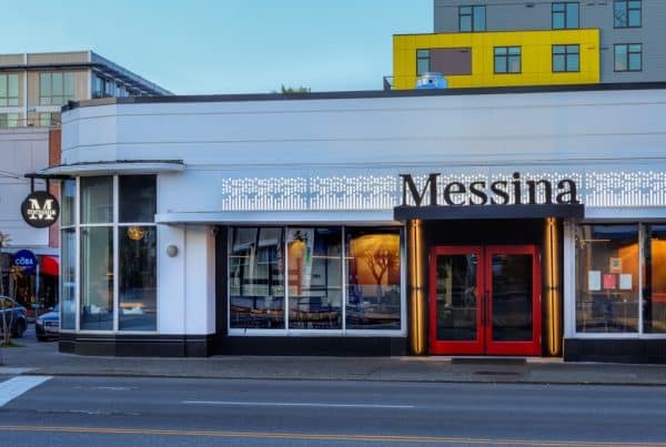 Street view of a modern building with a red door and the name "Messina" on the facade. Another distinct yellow building is visible in the background.