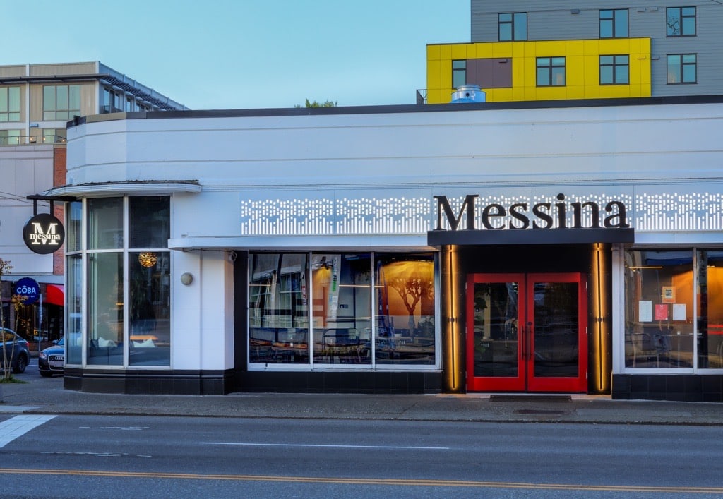 Street view of a modern building with a red door and the name "Messina" on the facade. Another distinct yellow building is visible in the background.