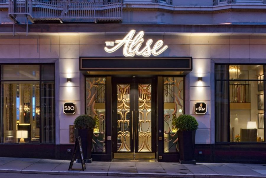 Street view of The Alise hotel entrance with illuminated sign, large windows on either side, potted plants, and a decorative glass door.