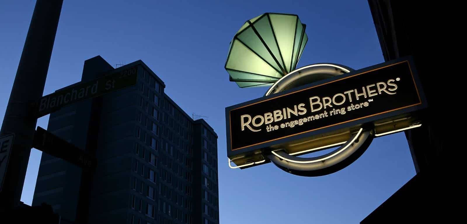 Illuminated sign for Robbins Brothers, the engagement ring store, against a backdrop of a city street with buildings at dusk.